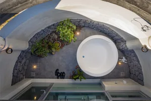 A modern bathroom featuring a circular bathtub surrounded by concrete tiles, plants, and black flip-flops.