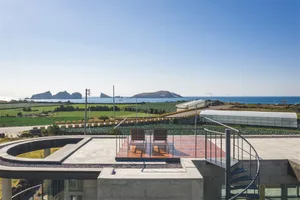 A modern residential exterior featuring a concrete patio with lounge chairs overlooking a lush green field and the ocean.