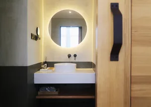 A minimalist bathroom with a circular mirror illuminated from behind, a white sink, and a wooden door.