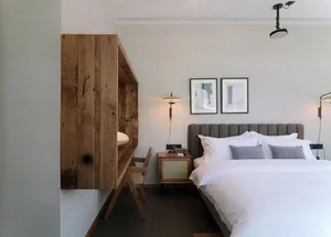 A minimalist bedroom featuring a wooden desk, concrete wall, and white textiles on a large bed.