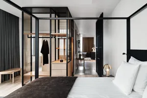 A minimalist bedroom with a bed, white bedding, and a glass-enclosed closet with wood shelves and metal accents.