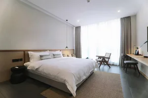 A minimalist bedroom with a large bed dressed in white linen, a wooden frame, and a hanging lamp above it.