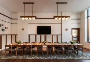 A modern dining room featuring a long, wooden dining table set for a large group, with pendant lights and a television in the