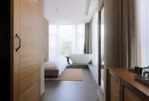 A minimalist bedroom with a wooden floor and concrete flooring, featuring a white bed and sheer curtains.