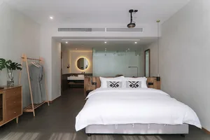 A minimalist bedroom featuring a white bed with geometric pillows, wooden headboard, and a glass partition in the background.