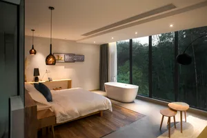 A modern minimalist bedroom featuring white bedding, wooden furniture, and a freestanding bathtub in front of a large window 