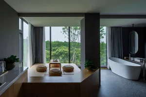 A modern residential bathroom with wood, concrete, and glass elements, featuring a bathtub, seating area, and large windows.