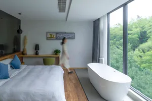 A modern bedroom with a white freestanding bathtub and a large window overlooking greenery.