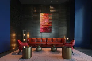 A modern lobby features a red leather sofa, two red chairs, and three metallic cylindrical tables on a patterned rug.