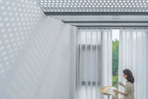 A woman reading a book near a window with sheer white curtains and a perforated ceiling.