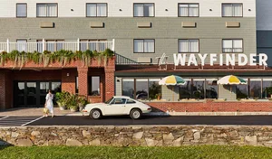A woman walks along the exterior of a modern hotel with a vintage white car parked nearby.