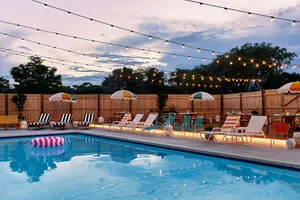 A serene residential backyard with a wooden fence, a sparkling swimming pool, colorful lounge chairs, and string lights.