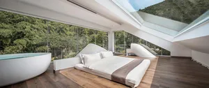 Modern minimalist bedroom with a bed, glass walls, and wooden flooring overlooking a forested area.