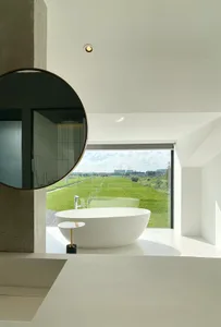 Modern bathroom with a minimalist white concrete bathtub, reflecting the lush greenery outside, and a large circular mirror.