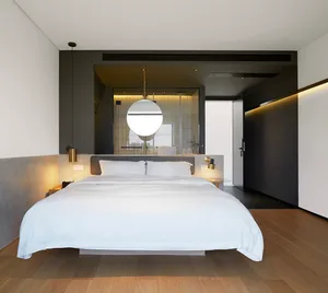A minimalist bedroom with a white bed on a concrete wall, wooden flooring, and a hanging light fixture.