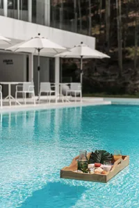 A wooden tray full of food floating in a pool, with white umbrellas and chairs in the background, creating a relaxing poolsid
