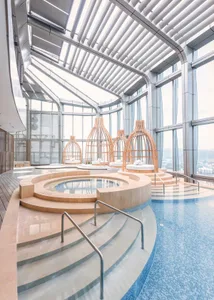 Modern swimming pool with glass walls and wooden cages, featuring plush seating and a view of the city.