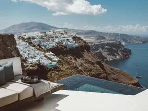 A luxurious outdoor living space with a modern infinity pool overlooking the ocean and a picturesque village on a cliff.
