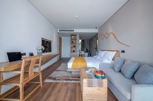 A modern hotel room with minimalist design features, including a wood desk, gray sofa, and a king-sized bed with white beddin