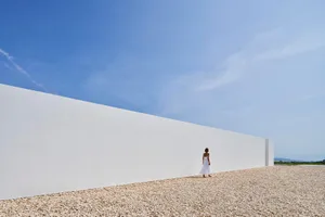 A minimalist white wall along a gravel ground with clear blue sky in the background.