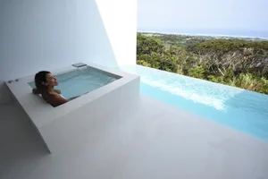 A woman relaxing in a minimalist concrete outdoor spa with scenic views of the forest and ocean.