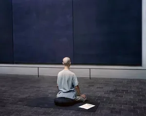 A minimalist installation art piece consisting of a person sitting on a cork mat and looking at a piece of paper in a museum 
