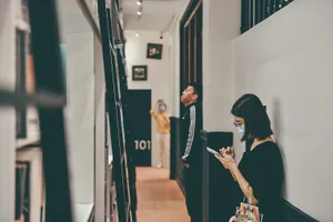 People standing in a narrow, well-lit corridor with a man wearing a yellow shirt in the background and a woman wearing a blac