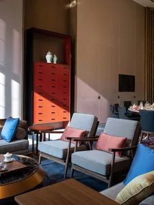 Modern interior design of a hotel lounge with grey armchairs, blue and pink cushions, and a wooden side table with a small te