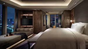 A contemporary bedroom featuring a large bed with white bedding, a seating area with a TV and lounge chairs, and a view of a 