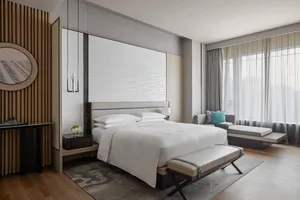 Modern, minimalist residential bedroom with white bedding, wooden floor, and large window.