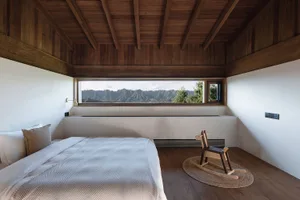 A minimalist bedroom with a large window overlooking mountains, featuring white walls, wood floors, a bed with a white comfor