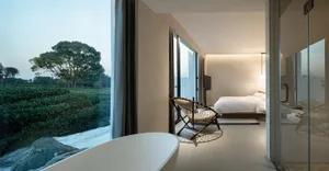 Modern minimalist bedroom with a wooden bed, glass walls, and fabric seating area overlooking greenery.