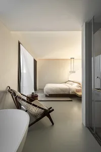 A minimalist bedroom featuring neutral-toned decor, white bedding, and a mid-century modern chair.