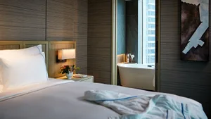 Modern bedroom with wooden headboard and bedside table, white bedding, and a bathtub with a city view.
