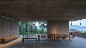A modern dining room with concrete walls and floor, featuring wooden tables and chairs, and a scenic outdoor view.