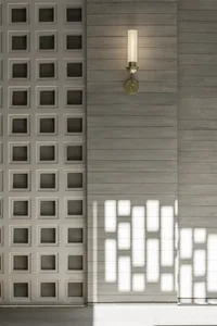A modern interior featuring a wall-mounted brass light fixture with a white lampshade, illuminated against a grey wooden pane