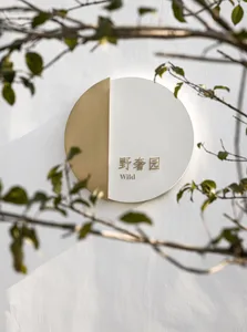 A modern, white and gold signage with Chinese characters and the word 'Wild' on it, mounted on a white wall with green foliag