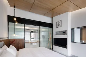 A minimalist bedroom featuring white bedding and lighting, with a glass-enclosed bathroom and wooden ceiling panels.