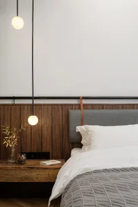 A minimalist bedroom featuring a wooden wall, a bed with a grey headboard, and a hanging light fixture.
