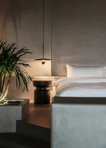 A modern bedroom with a minimalist design, featuring a concrete headboard and flooring, and a hanging pendant light.