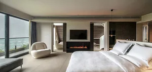 Modern minimalist bedroom featuring a large bed with white bedding, a glass fireplace, and large windows with glass walls.