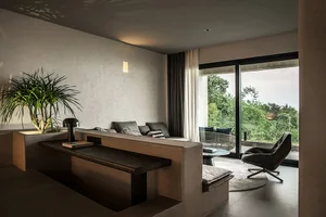 A minimalist living room with concrete walls, a gray sofa, and a black armchair, featuring a large window with a view of gree