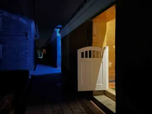 A white wooden gate illuminated by a warm glow, contrasting with the dark surroundings, leading into a warmly lit interior.