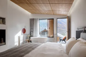 A minimalist bedroom featuring a bed with white bedding, a freestanding bathtub, and a view of mountains through a window wit