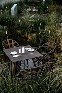 Modern outdoor dining area with metal chairs and glass table surrounded by tall grasses and illuminated by string lights.