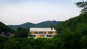 Modern house with white walls surrounded by green trees and mountains in the background.