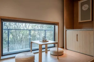 A modern tea room featuring woven stools, a wooden table, and a large window overlooking the trees.
