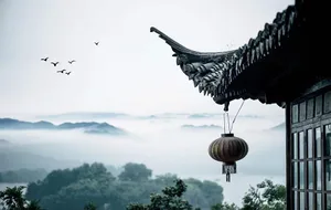 A ceramic lantern hangs from the eaves of a traditional-style building, with a scenic landscape of misty mountains and flying