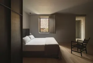 A minimalist bedroom with a bed, chair, and small window overlooking a tiled rooftop.