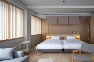 Minimalist hotel room with twin beds, wood furniture, cloth wall coverings, and light fixtures.